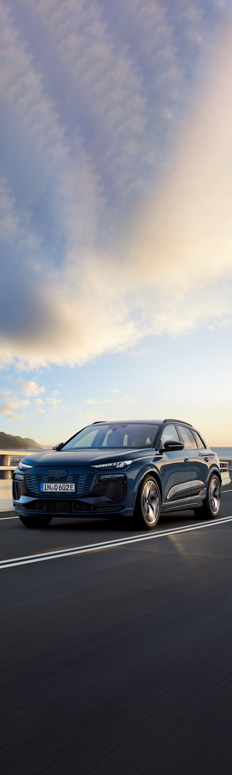 Three-quarter front view of the Audi Q6 e-tron in motion on a coastal road.